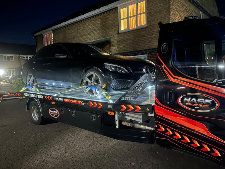 Black car loaded on a Hass Recovery tow truck at night, with illuminated house in background